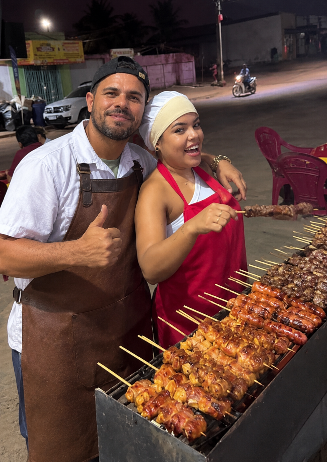 Marconi e Camila atrás da churrasqueira cheia de espetinhos, à noite